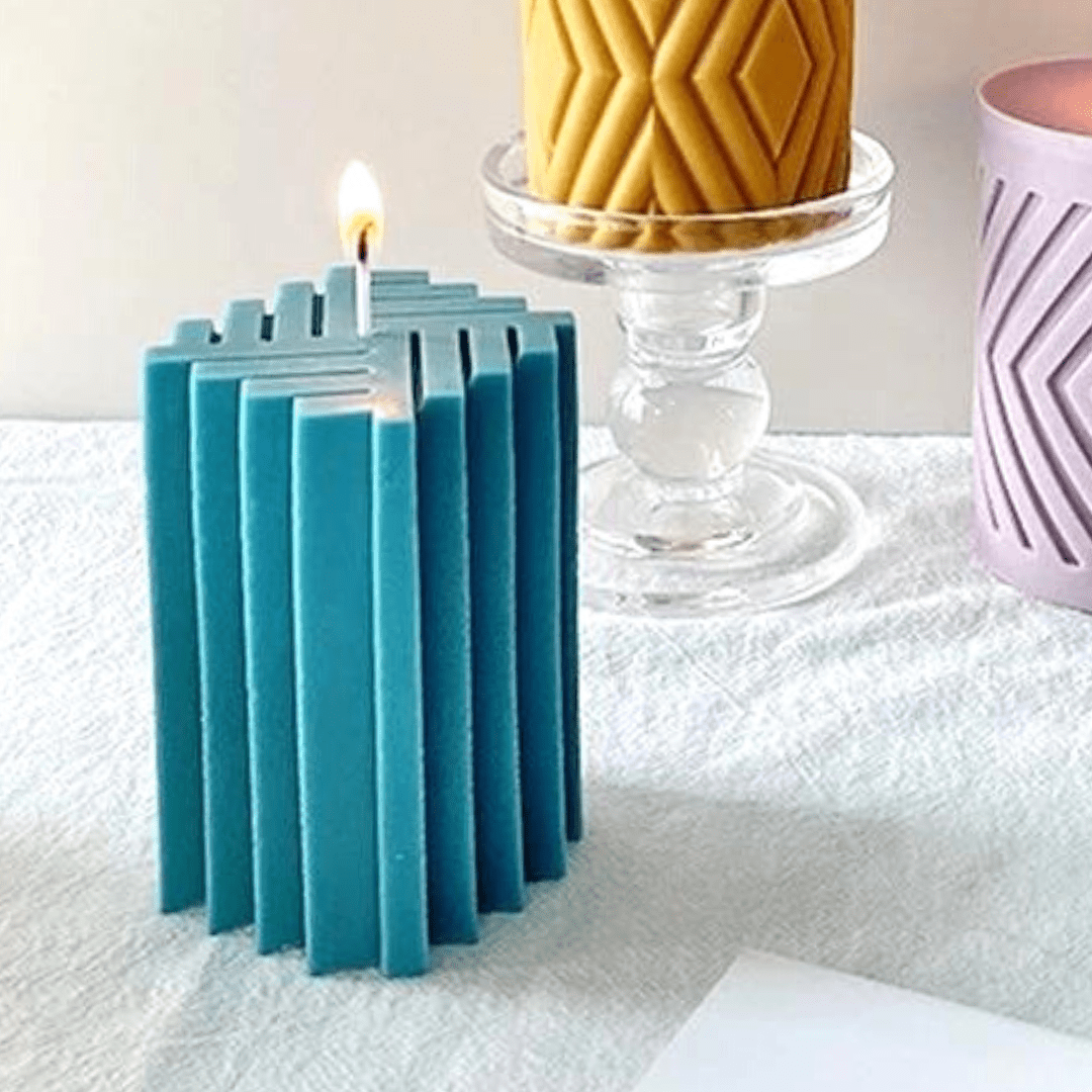 blue geometric square shaped candle with wick and flame on textured white table cloth with various decor items in background