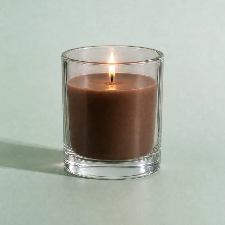 Pure Candle Supplies Melbourne dark brown candle in a clear glass container with a lit wick on a light green surface.