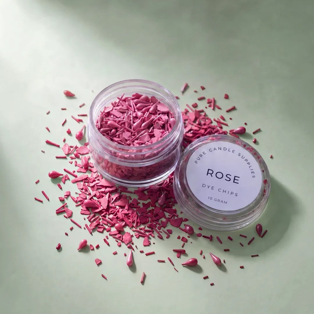 Pure Candle Supplies Melbourne rose dye chips in a small clear jar with scattered pink wax pieces on a green surface.