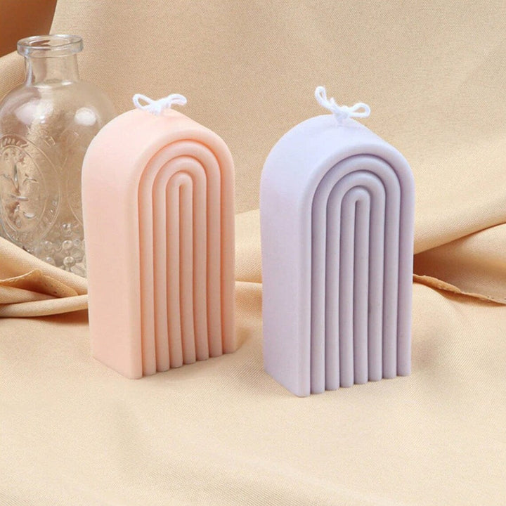 two tall rainbow shaped candles in peach and purple colours on a beige background with a glass bottle in the background
