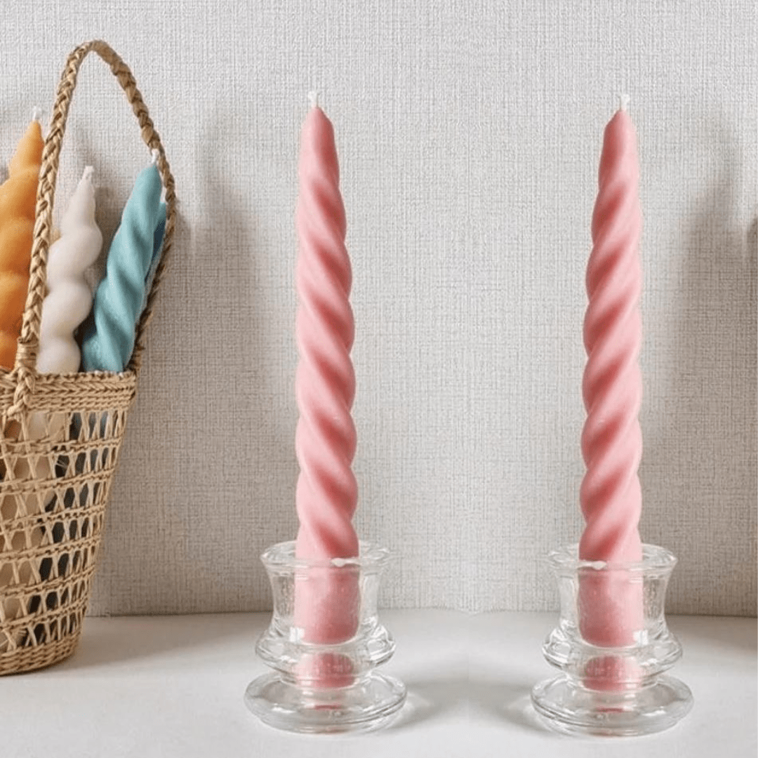 two pink spiral candlesticks in glass holders against a beige linen wall with a basket of spiral candlesticks in various colours in background